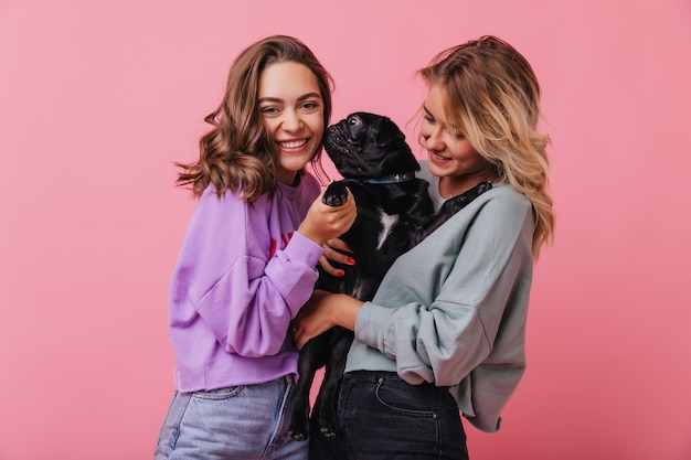 Incantevole ragazza dai capelli castani che tocca un simpatico cucciolo nero Elegante donna bionda in abito casual che tiene il suo cane su sfondo rosa