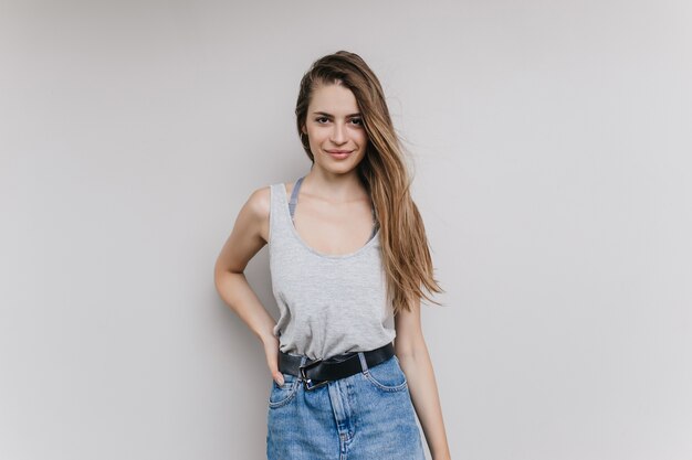 Incantevole ragazza dai capelli lunghi in posa con l'espressione del viso soddisfatto. Ritratto dell'interno della giovane signora castana sicura in gonna del denim.