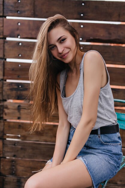 Incantevole ragazza europea in breve gonna di jeans in posa sulla parete in legno. Carina giovane signora con sorriso timido godendo la giornata di primavera.