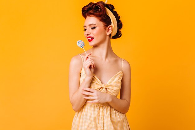 Incantevole ragazza pinup con caramelle dure. Studio shot di pensieroso donna allo zenzero con lecca-lecca isolato su spazio giallo.