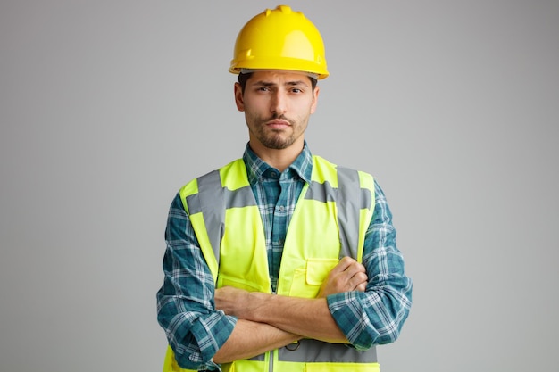 Incerto giovane ingegnere maschio che indossa casco di sicurezza e uniforme guardando la fotocamera tenendo le braccia incrociate isolate su sfondo bianco