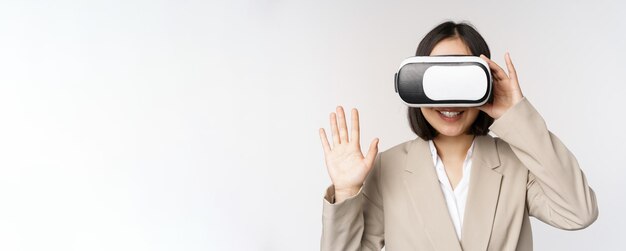 Incontro in chat vr Donna d'affari asiatica con occhiali per realtà virtuale alzando la mano e salutando qualcuno in piedi su sfondo bianco
