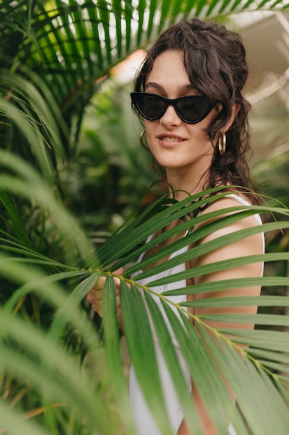 Incredibile bella donna alla moda in occhiali da sole con bei guadagni in posa attraverso alberi esotici