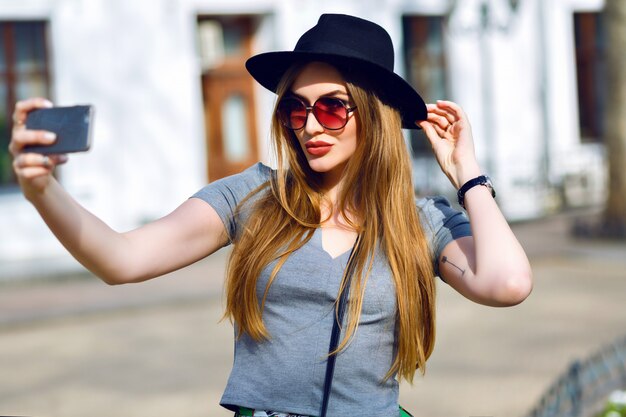 Incredibile donna bionda che fa selfie per strada, cappello hipster e occhiali da sole, labbra carnose e capelli lunghi, vacanze in Europa.