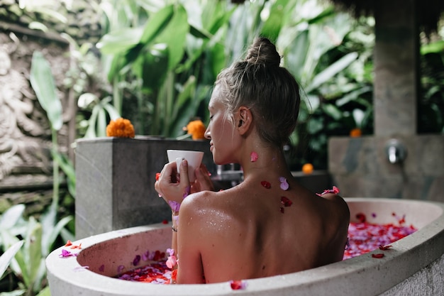 incredibile donna che beve il tè con gli occhi chiusi mentre era seduto in bagno. Ritratto dal retro della signora allegra con pelle abbronzata facendo spa con petali di rosa.