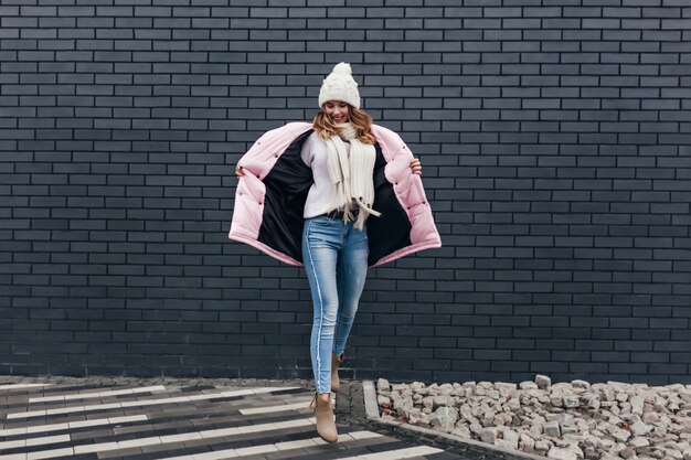 Incredibile donna in abbigliamento invernale divertente ballare sulla strada urbana. Modello femminile in jeans che gode del servizio fotografico in una giornata fredda.