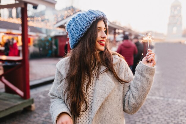 Incredibile donna in cappotto grigio e cappello blu che cammina per strada con sparkler. Adorabile donna femmina in abito invernale trascorrere del tempo all'aperto e guardando la luce del Bengala con un sorriso.