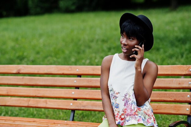 Incredibile donna modello afroamericano in pantaloni verdi e cappello nero in posa su una panchina al parco e parlando al telefono cellulare