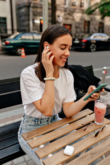 Incredibile donna tenera con un sorriso felice che indossa una maglietta bianca con in mano le cuffie wireless e guarda lo smartphone Bella signora che ascolta musica in cuffia e beve un frullato estivo all'esterno