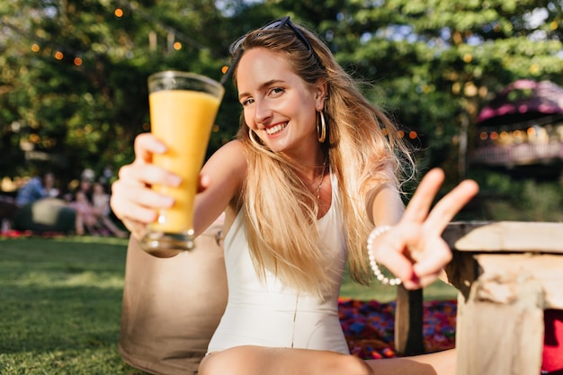 Incredibile giovane donna con un sorriso carino in posa con un bicchiere di succo d'arancia sullo sfondo della natura Bella ragazza bionda in abito bianco che gode di un cocktail di frutta nel parco nella mattina d'estate