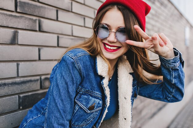 Incredibile ragazza bianca in occhiali rotondi alla moda in posa con il segno di pace. Colpo esterno di donna bruna intelligente che ride nel muro di mattoni di sfocatura.