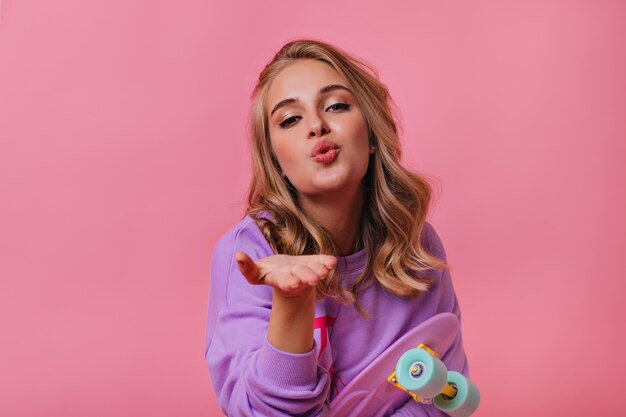 Incredibile ragazza bionda con trucco alla moda in posa con lo skateboard. Adorabile signora caucasica che invia un bacio d'aria su pastello.