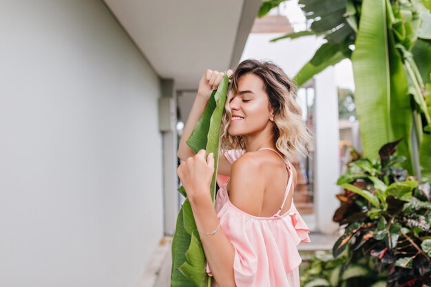 Incredibile ragazza con taglio di capelli carino in posa con gli occhi chiusi vicino al giardino. Foto all'aperto di estatica donna abbronzata in abito rosa alla moda.