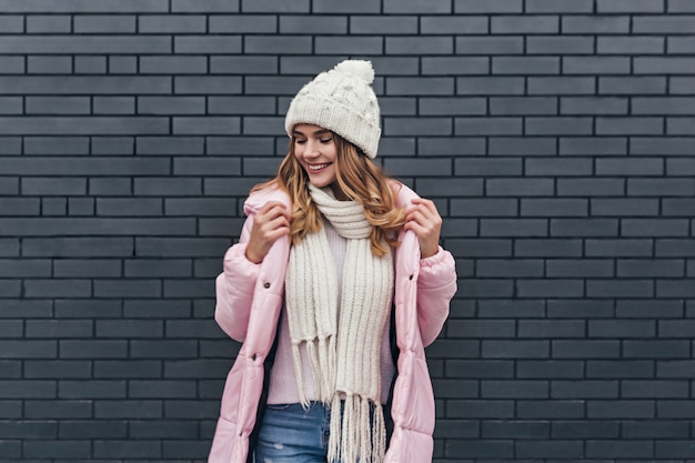 Incredibile ragazza con un bel sorriso che gode del servizio fotografico nella fredda mattina. Modello femminile biondo alla moda in cappotto e cappello lavorato a maglia in posa vicino al muro di mattoni.