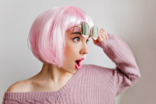 Incredibile ragazza in peruke rosa in posa con stupore e distogliere lo sguardo. Affascinante modello femminile in parrucca colorata in piedi sulla parete chiara con gli occhiali.