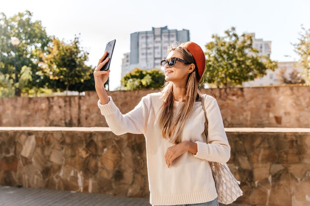 Incredibile signora francese in camicia bianca che fa selfie nel fine settimana autunnale. Adorabile ragazza splendida in berretto rosso in piedi sulla strada con il telefono.