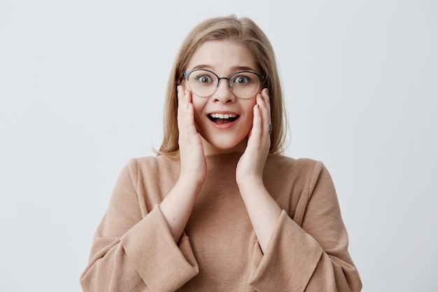 È incredibile! Sorpresa donna emotiva sbalordita tiene entrambe le mani sulle guance, scioccata dal vedere i prezzi elevati, isolata contro il muro bianco dello studio. La femmina bionda non può credere in notizie scioccanti