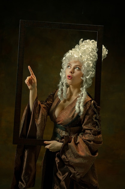 Indicando. Ritratto di giovane donna medievale in abiti vintage con cornice in legno su sfondo scuro. Modello femminile come duchessa, persona reale. Concetto di confronto di epoche, moderno, moda, bellezza.
