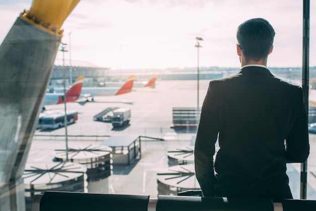 Indietro di un giovane uomo d'affari in piedi con la valigia in aeroporto in attesa del volo xA