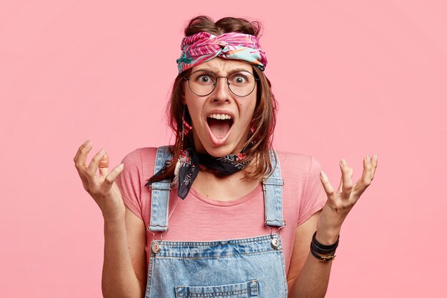 Indignata donna hippy arrabbiata gesticola con le mani, esclama furiosamente, esprime emozioni negative, vestita con tuta e fascia alla moda, posa contro il muro rosa