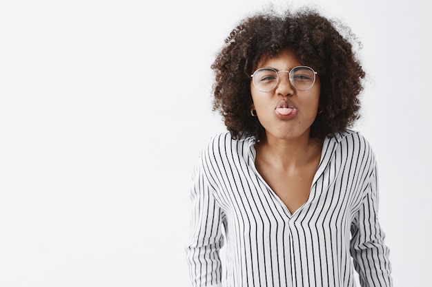 infantile e giocoso divertente afroamericano donna adulta con acconciatura afro in ufficio a strisce camicetta e bicchieri piegandosi verso e mostrando la lingua rughe naso con gioia