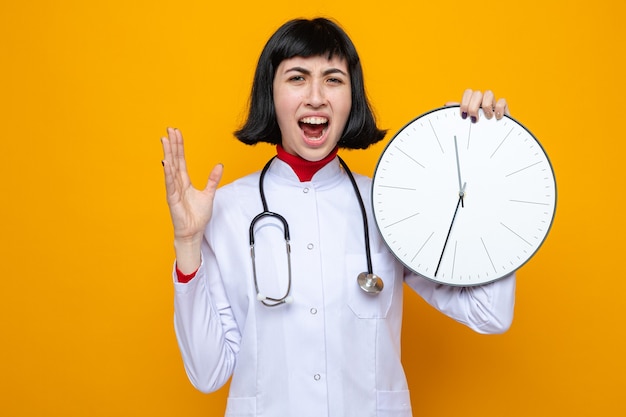 Infastidita giovane bella ragazza caucasica in uniforme da medico con lo stetoscopio che tiene l'orologio e tiene la mano aperta