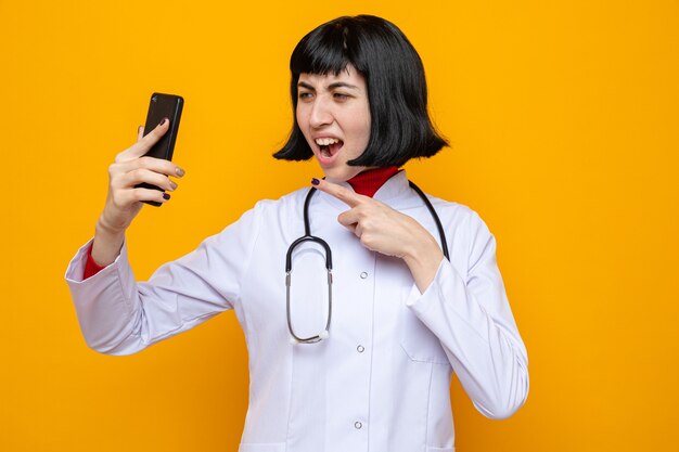 Infastidita giovane bella ragazza caucasica in uniforme da medico con stetoscopio che tiene in mano e punta al telefono