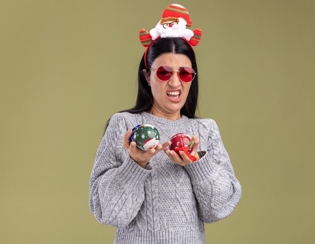 Infastidita giovane ragazza caucasica indossando la fascia di Babbo Natale con gli occhiali che tengono le bagattelle di Natale che guarda l'obbiettivo isolato su sfondo verde oliva