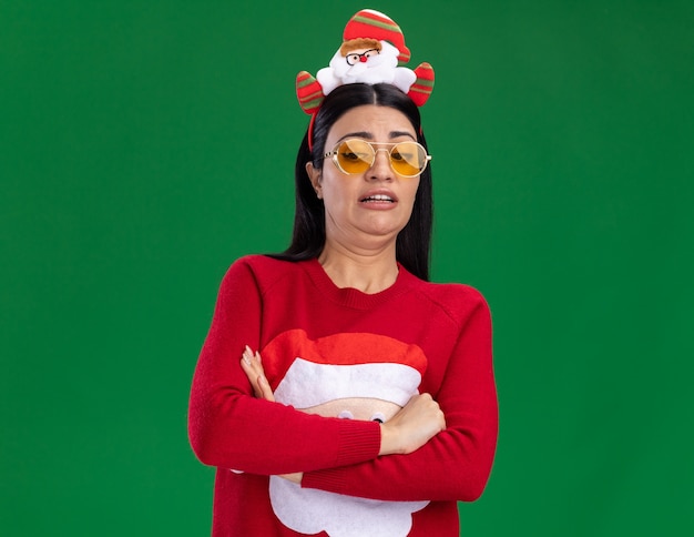 Infastidita giovane ragazza caucasica indossando la fascia di Babbo Natale e maglione con gli occhiali in piedi con la postura chiusa guardando verso il basso isolato sulla parete verde con spazio di copia