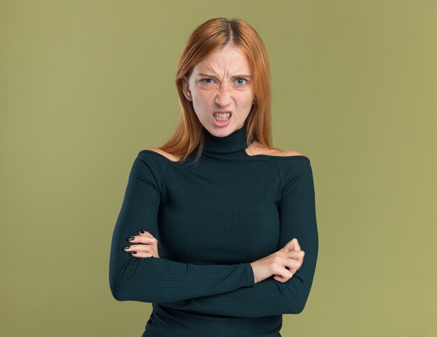 Infastidita giovane ragazza rossa allo zenzero con le lentiggini in piedi con le braccia incrociate isolate sulla parete verde oliva con spazio di copia