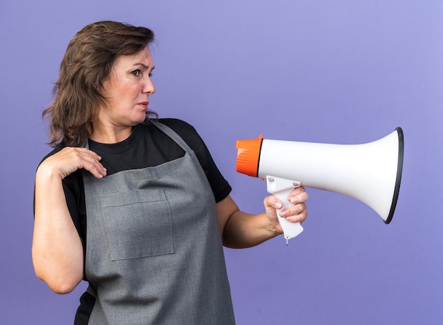 Infastidito adulto femmina caucasica barbiere in uniforme tenendo altoparlante e guardando il lato isolato su sfondo viola con spazio di copia