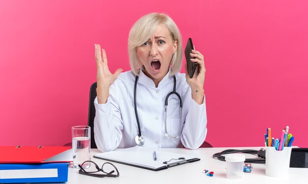 infastidito dottoressa adulta in veste medica con stetoscopio seduto alla scrivania con strumenti da ufficio tenendo il telefono e urlando a qualcuno isolato sulla parete rosa con spazio di copia