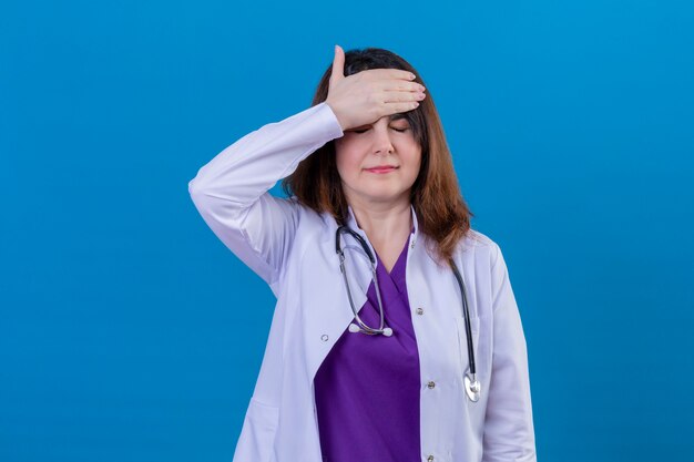 Infastidito e infastidito donna di mezza età medico indossa camice bianco e con lo stetoscopio in piedi con la mano sulla testa sopra lo spazio blu