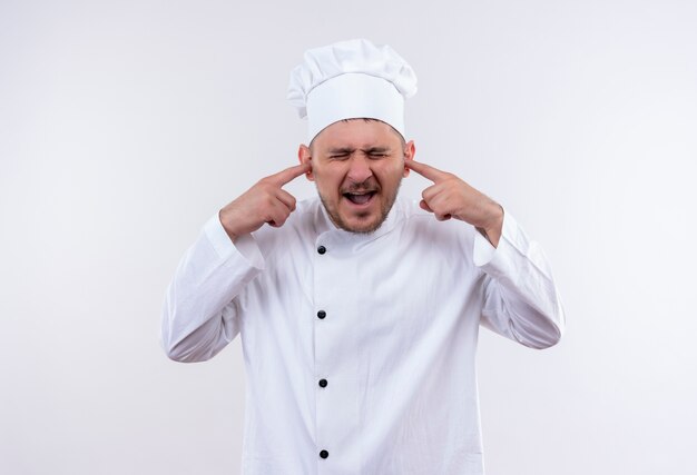 Infastidito giovane bel cuoco in uniforme da chef che mette le dita nelle orecchie con gli occhi chiusi isolati sul muro bianco white