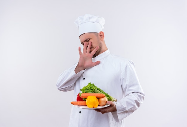 Infastidito giovane bel cuoco in uniforme da chef tenendo piatto con verdure e gesticolando no sul muro bianco isolato