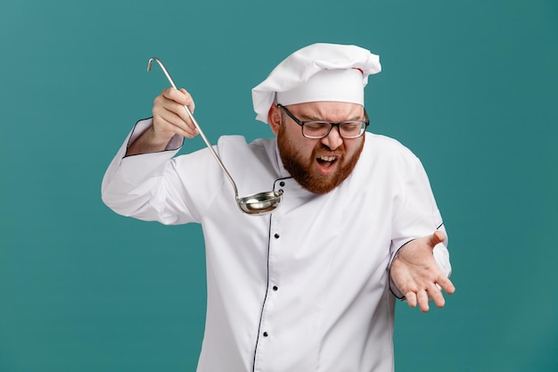 Infastidito giovane chef maschio che indossa occhiali uniforme e cappuccio che tiene la siviera guardando in basso mostrando la mano vuota isolata su sfondo blu