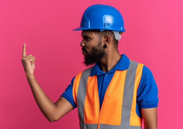 Infastidito giovane costruttore afro-americano uomo in uniforme con casco di sicurezza guardando il dito isolato su sfondo rosa con copia spazio