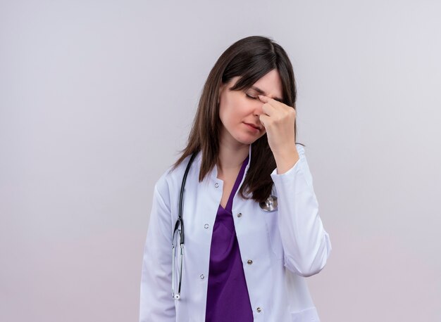 Infastidito giovane dottoressa in abito medico con lo stetoscopio mette la mano sul viso isolato su sfondo bianco con copia spazio