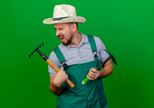 Infastidito giovane giardiniere slavo bello in uniforme e cappello che tiene zappa-rastrello e rastrello guardando rastrello