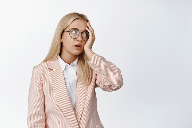 Infastidito giovane impiegato femminile, occhi al cielo e facepalm stanco, irritato con qualcosa, in piedi in tuta e occhiali su bianco.