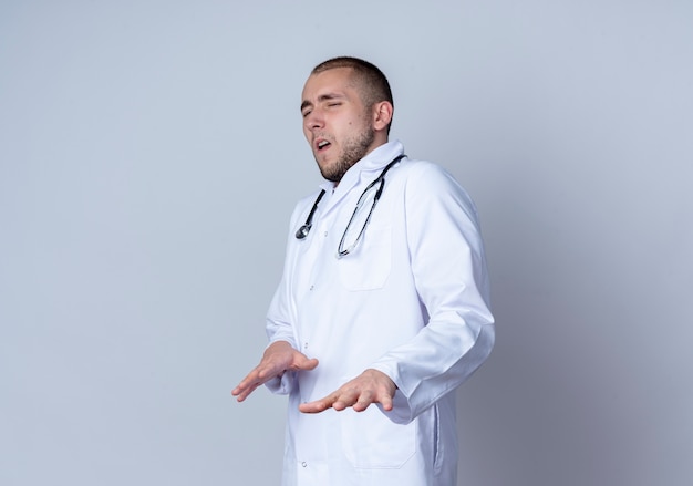 Infastidito giovane maschio medico indossando abito medico e stetoscopio intorno al collo in piedi in vista di profilo allungando le mani isolate su bianco con spazio di copia