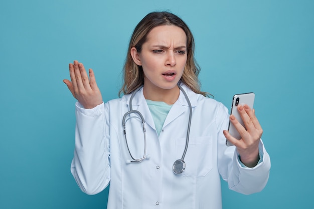 Infastidito giovane medico femminile che indossa abito medico e stetoscopio intorno al collo tenendo e guardando il telefono cellulare tenendo la mano in aria