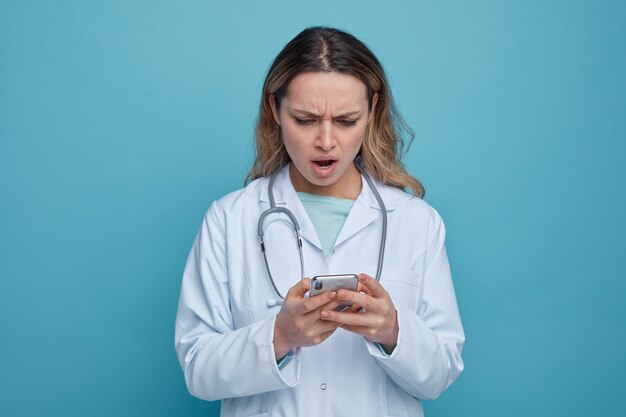 Infastidito giovane medico femminile che indossa abito medico e stetoscopio intorno al collo utilizzando il suo telefono cellulare