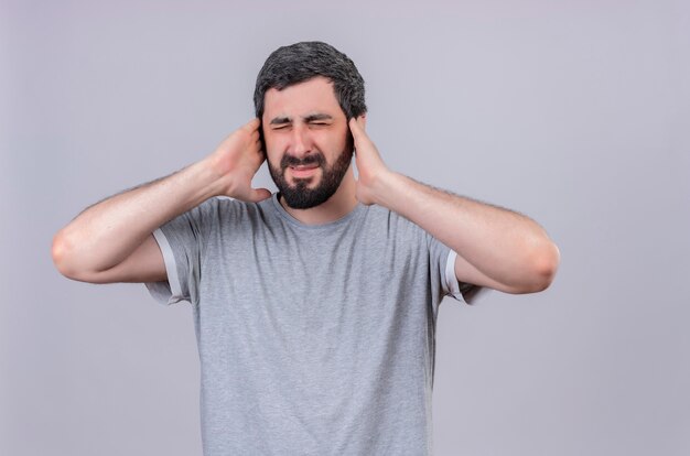 Infastidito giovane uomo caucasico bello che mette le mani sulle orecchie con gli occhi chiusi isolati su bianco