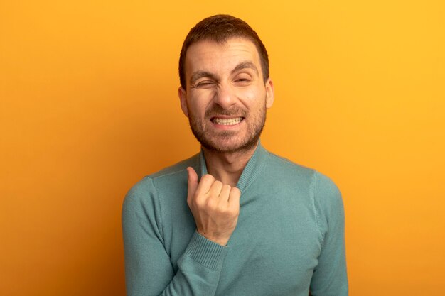 Infastidito giovane uomo caucasico tirando il collare del suo maglione a collo alto guardando la fotocamera con un occhio chiuso isolato su sfondo arancione con spazio di copia