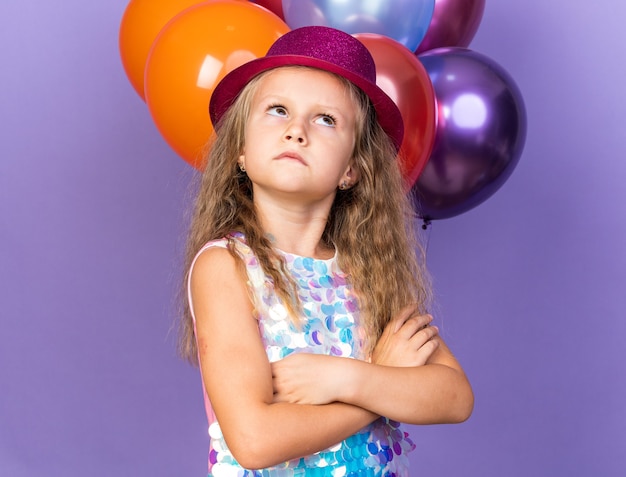 infastidito, piccola ragazza bionda con viola party hat in piedi con le braccia incrociate davanti a palloncini di elio cercando isolato sulla parete viola con spazio di copia