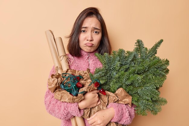 Infelice donna bruna cupa con aspetto orientale tiene oggetti per le vacanze per la decorazione si prepara per la celebrazione del nuovo anno indossa cappotto invernale isolato su sfondo beige. Concetto della vigilia di Natale