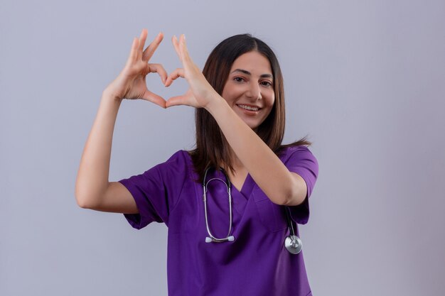 infermiera che indossa l'uniforme e lo stetoscopio che fa un gesto romantico del cuore con il sorriso sul viso in piedi sul bianco isolato