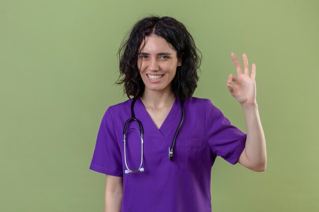 infermiera che indossa l'uniforme e lo stetoscopio sorridendo allegramente facendo segno ok con la faccia felice in piedi sul verde isolato