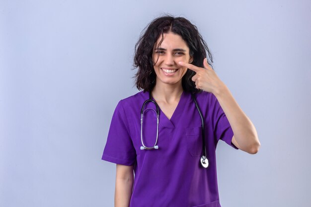 Infermiera della giovane donna in uniforme medica e con lo stetoscopio che sorride con la faccia felice che indica con il dito alla sua condizione del naso